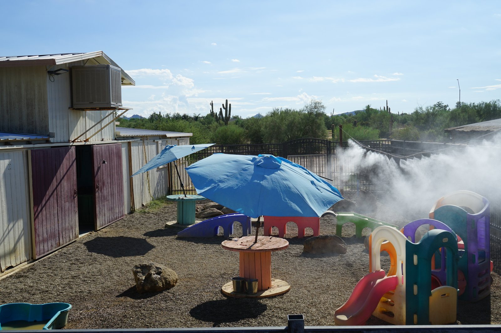 The Cave Creek desert play yard with overhead misters spraying cooling mist over the gravel, a blue umbrella and play structures, built for beating the heat.