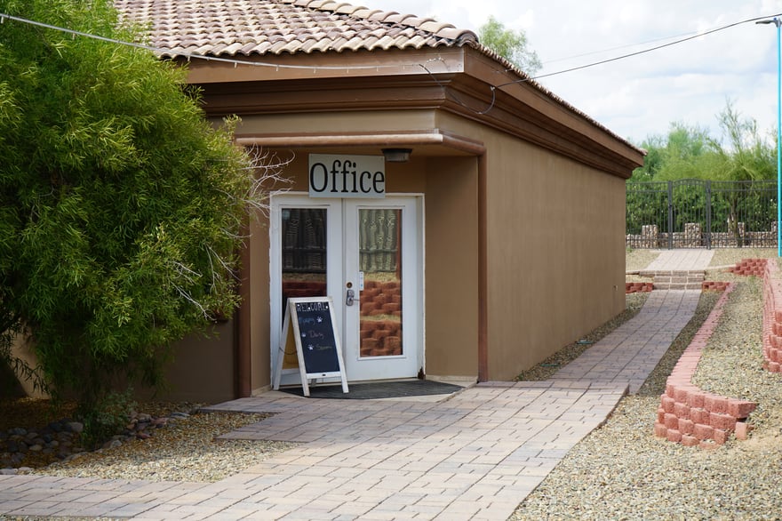 Exterior of the tan stucco Partners Dogs facility with glass Office doors and a paver walkway, an inviting establishing shot for the contact CTA.