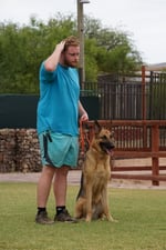 Partners Dogs trainer in a teal shirt standing beside a German Shepherd holding a calm sit-stay during an outdoor obedience training session
