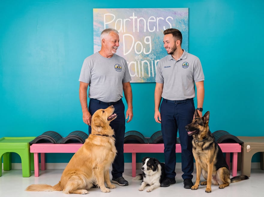 Two friendly Partners Dog Training team members smiling beside the branded sign, ready to help you find the right plan.
